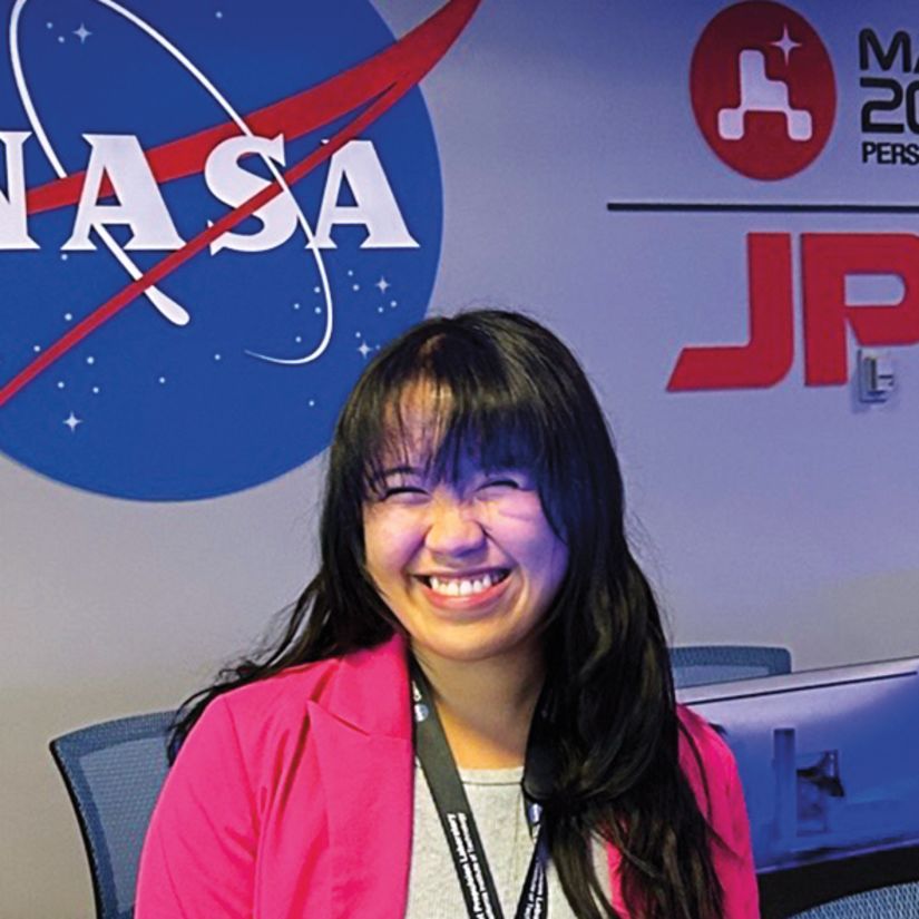 Joan Marie Tubungbunua is wearing a bright pink blazer and smiling widely. She’s in the middle of a control room where many chairs and computer monitors are lined up in two rows.