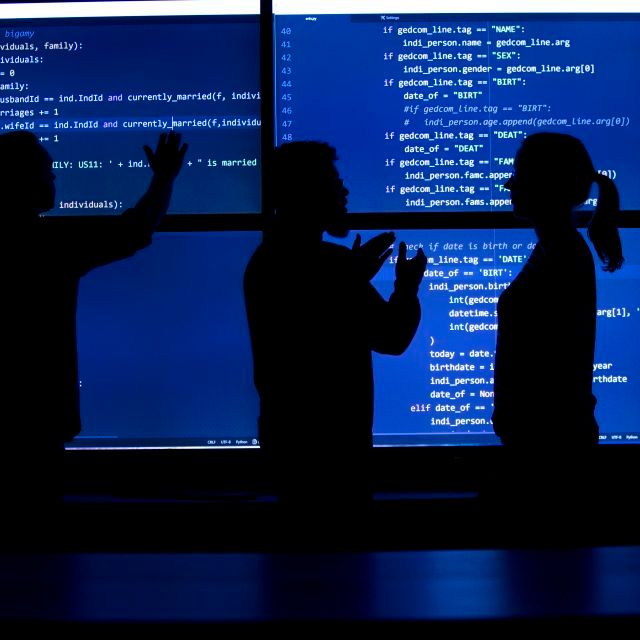 A silhouette of three people looking at a blue screen in a dark room.