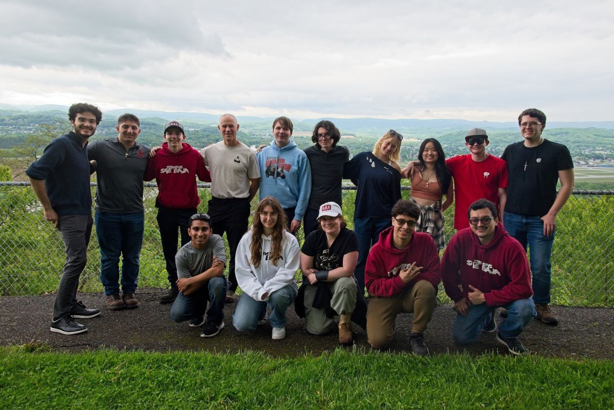 Two rows of people stand (back row) and kneel (front row) outdoord in front of a chain-linked fence with a green landscape view in the background