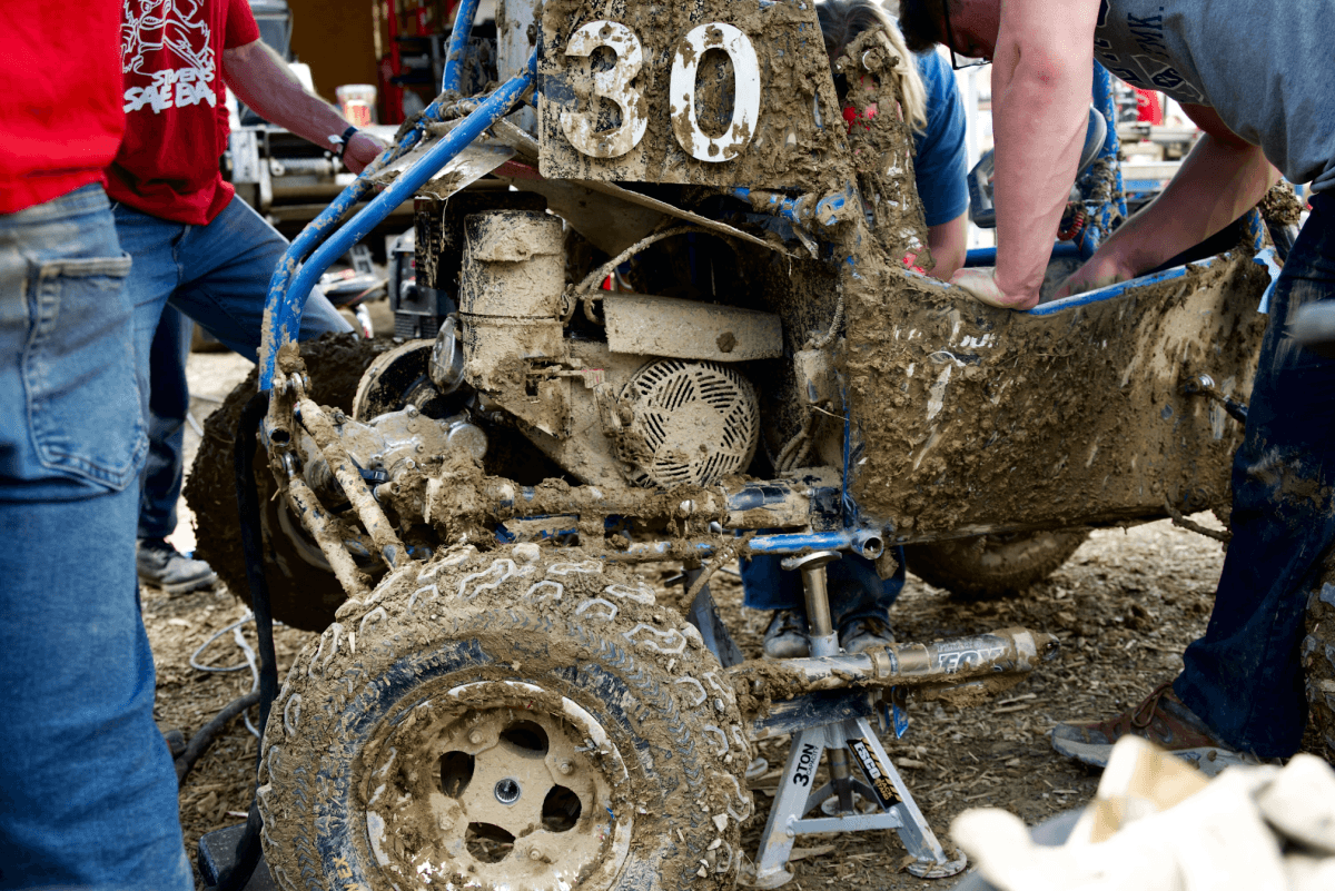 A close-up of a muddy off-road vehicle and a wheel with the arms and legs of some people working on it