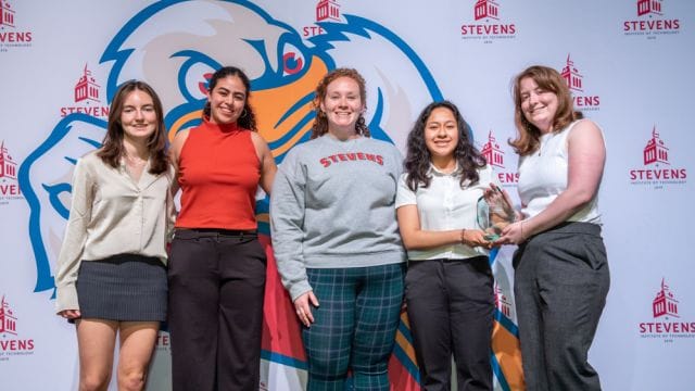 A photo of 5 young women standing together and holding an award