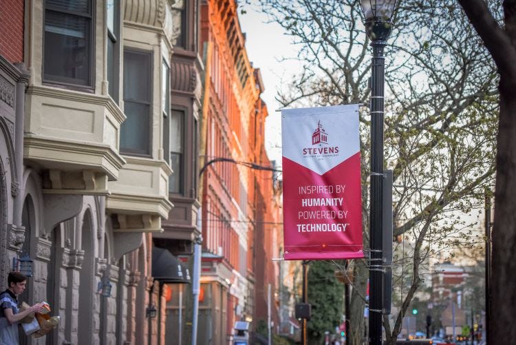 A Stevens banner saying Inspired by Humanity, Powered by Technology on a streetlight along a Hoboken Street.