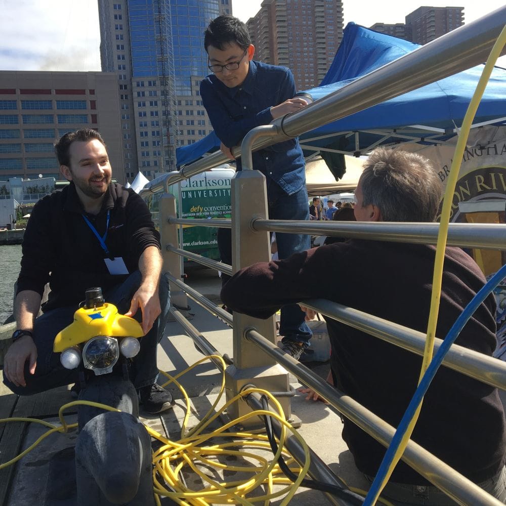 Stevens professor Brendan Englot and two onlookers on a pier with Englat's autonomous water robot.