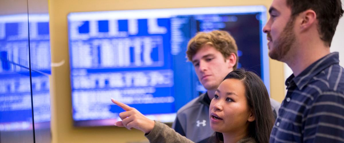 Student points at wall monitor while two other students look on.