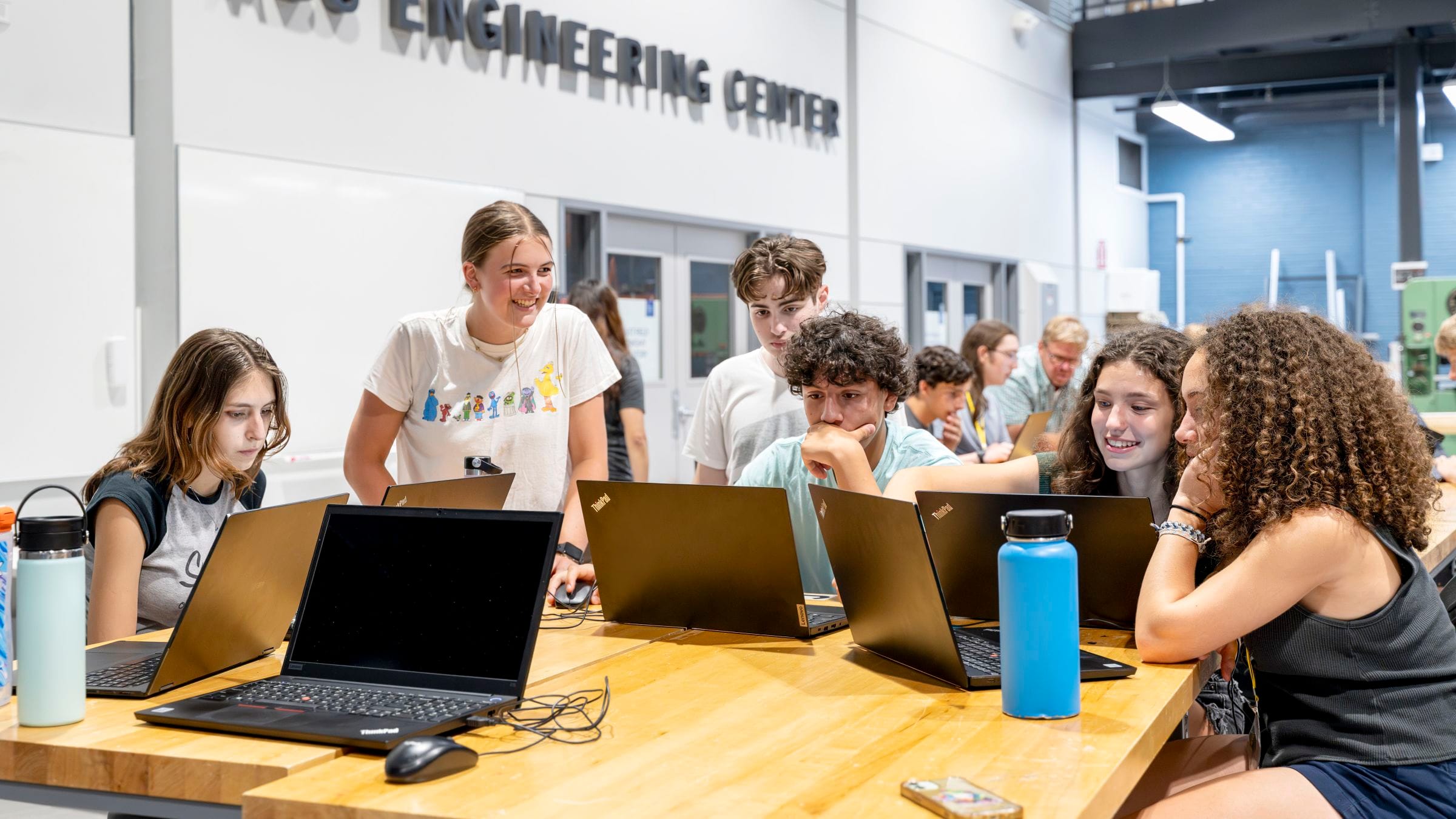 A group of six high school students, two boys and four girls, sit at a table in front of laptops, discussing a project in the ABS Engineering Center. 