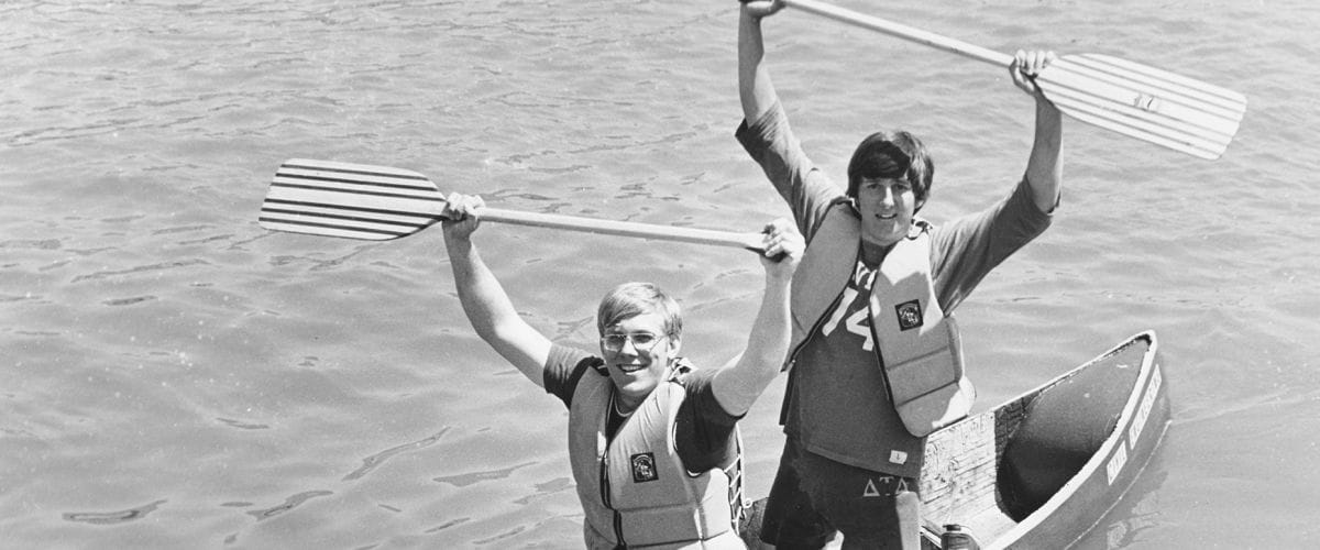 Two young men are in a canoe, one behind the other. The man in front is sitting, holding an oar in both hands over his head. The man in back is kneeling, also holding an oar in both hands over his head.