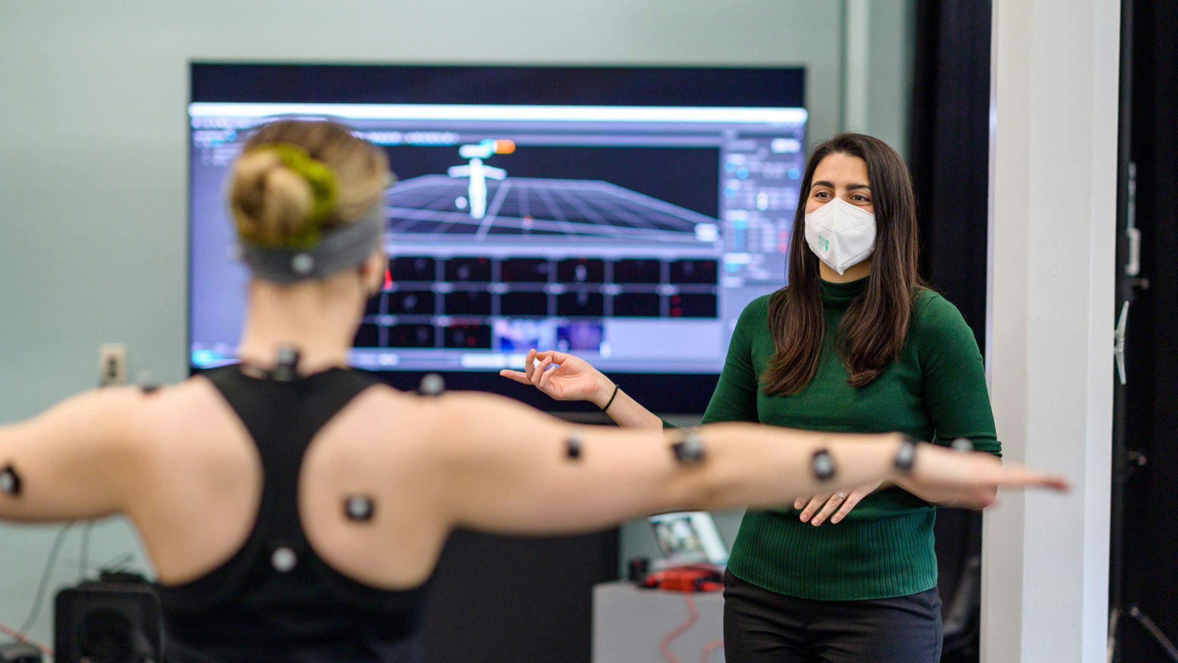 Model with motion capture nodes attached to arms raises arms with instructor in background