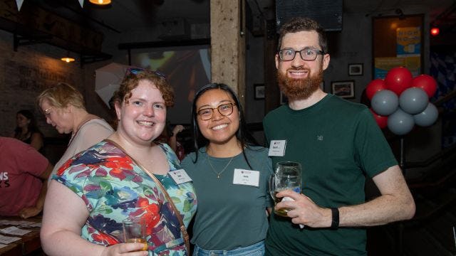Three young alumni hold drinks and smile at event