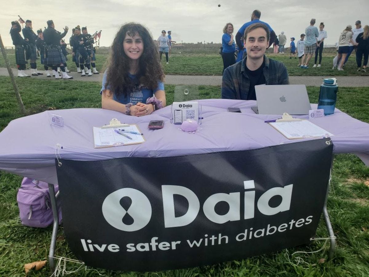 Arianna Gehan ’24 and Frank Pinnola ’23 sit outdoors at a table with a banner that reads "Daia - live safer with diabetes"