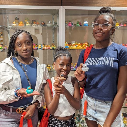 Three young women holding rubber ducks in front of a glass display case containing more ducks.