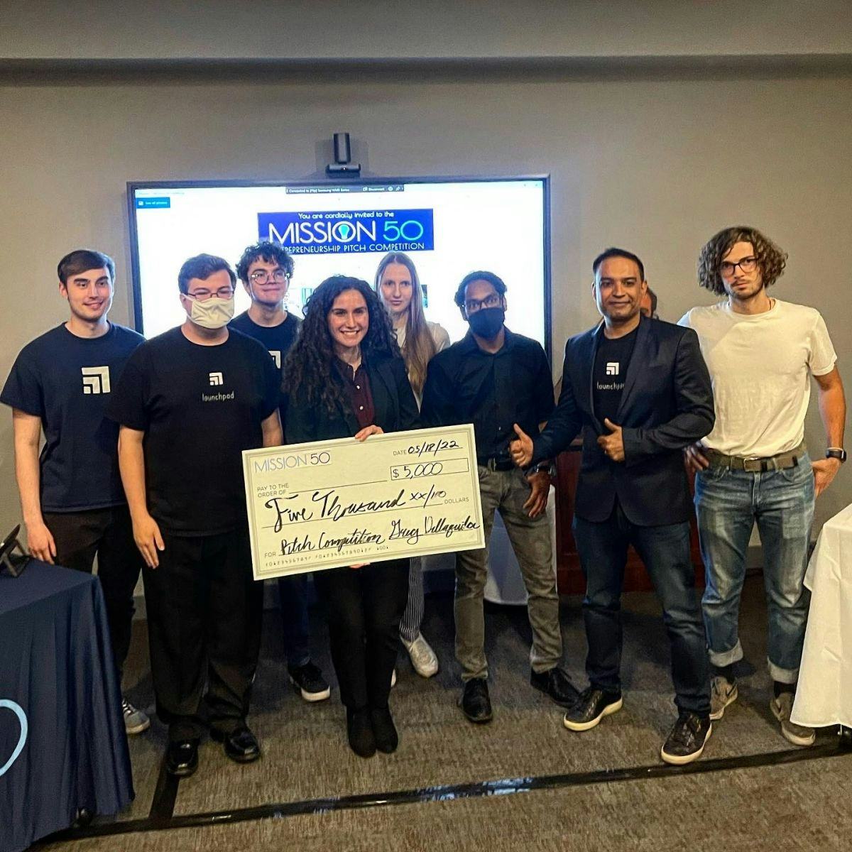 Arianna Gehan holds giant check while standing in a group in front of a sign that reads "Mission 50 Entrepreneurship Pitch Competition." The check is written from Mission 50 for $5000.
