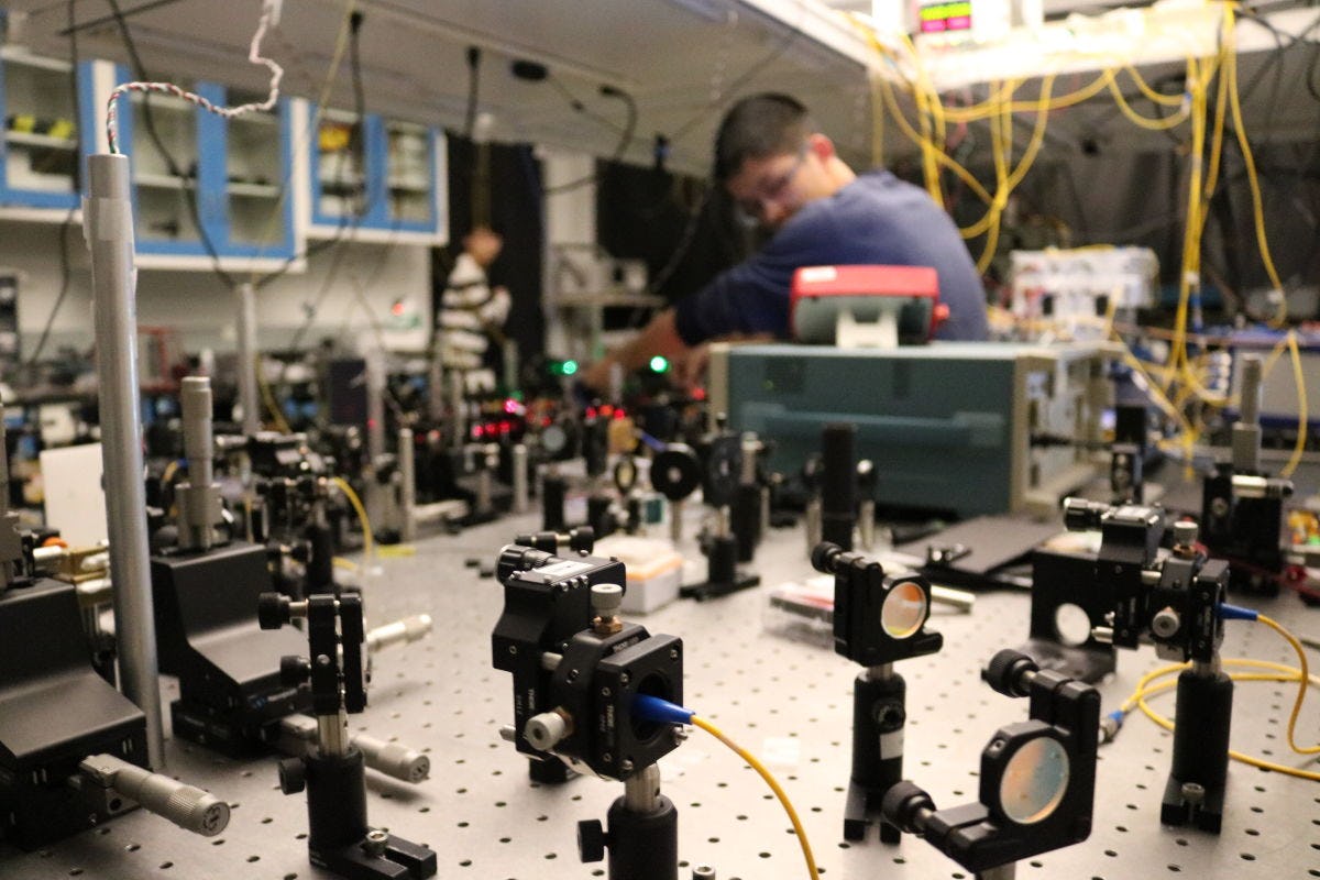 A photo of a male student working in the quantum lab surrounded by wires and photonics equipment.
